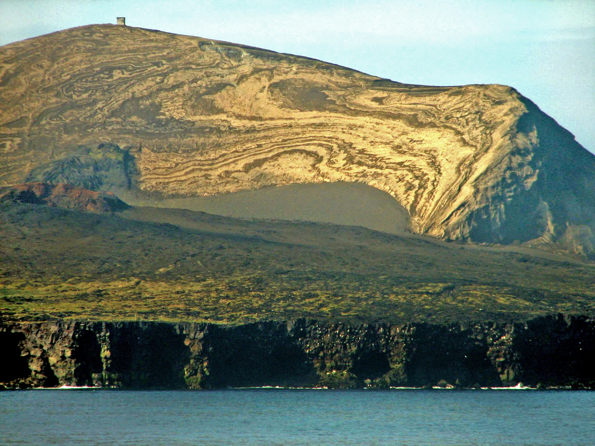 A wide view of the volcanic island of Surtsey in Iceland, featuring layered sediment slopes, dark cliffs, and a small structure atop the summit.