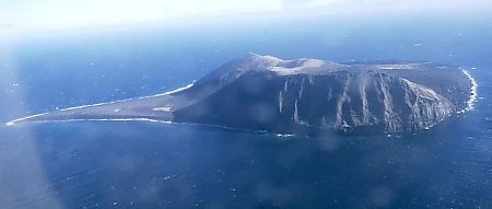 Aerial view of the isolated, rocky Surtsey island surrounded by deep blue ocean, featuring a volcanic cone and sloped terrain.