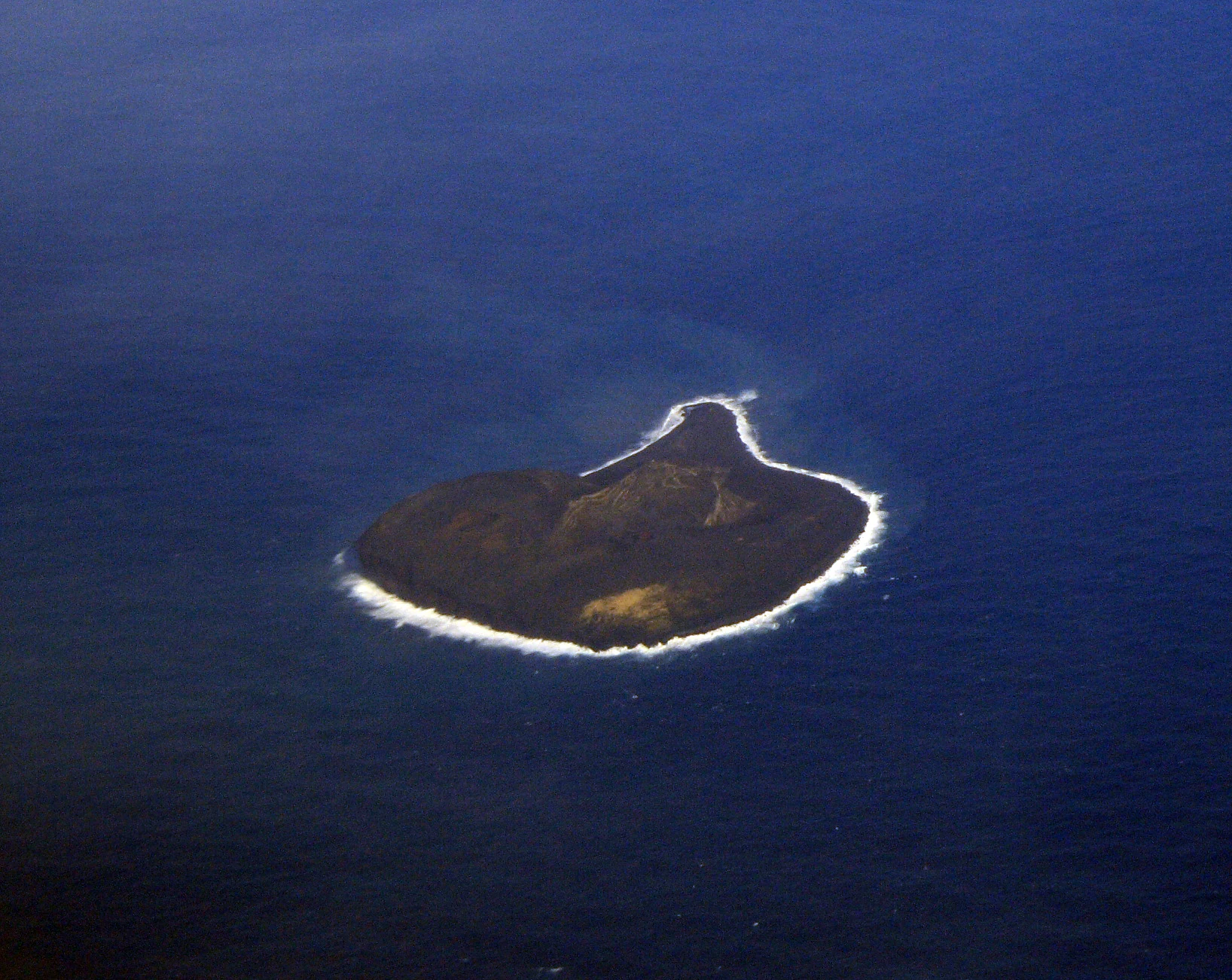 An aerial view of the small, dark, volcanic island of Surtsey surrounded by deep blue ocean.