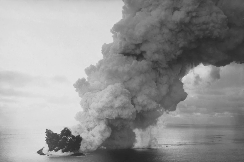 Black and white photograph showing a volcanic eruption occurring at sea; a large, dark island mass is visible with a massive plume of white smoke and ash rising into the sky.