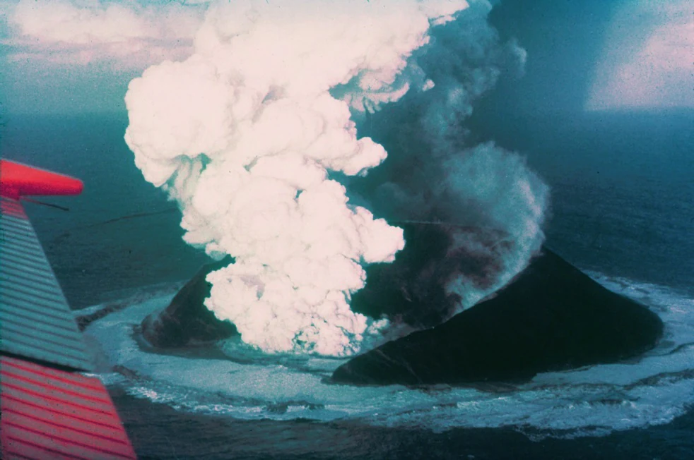 Aerial view looking down at a volcanic eruption occurring in the ocean, with plumes of white steam and dark ash rising from the water, and the wing of an aircraft visible in the foreground.