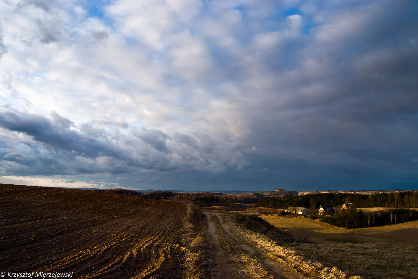 Una toma amplia y a baja altura de un paisaje rural con un camino de tierra en primer plano, colinas onduladas y un cielo azul dramático y nublado.