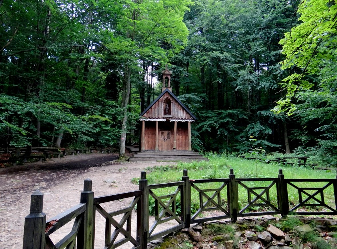 Una pequeña capilla de madera con techo a dos aguas se alza en un claro de bosque frondoso y verde, vista detrás de una valla de madera baja.