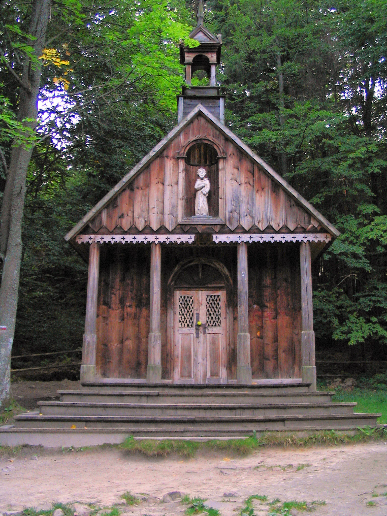 Una vista frontal de una pequeña capilla o ermita de madera tradicional, enclavada en un entorno forestal, con una escultura de un santo en un nicho sobre la entrada.