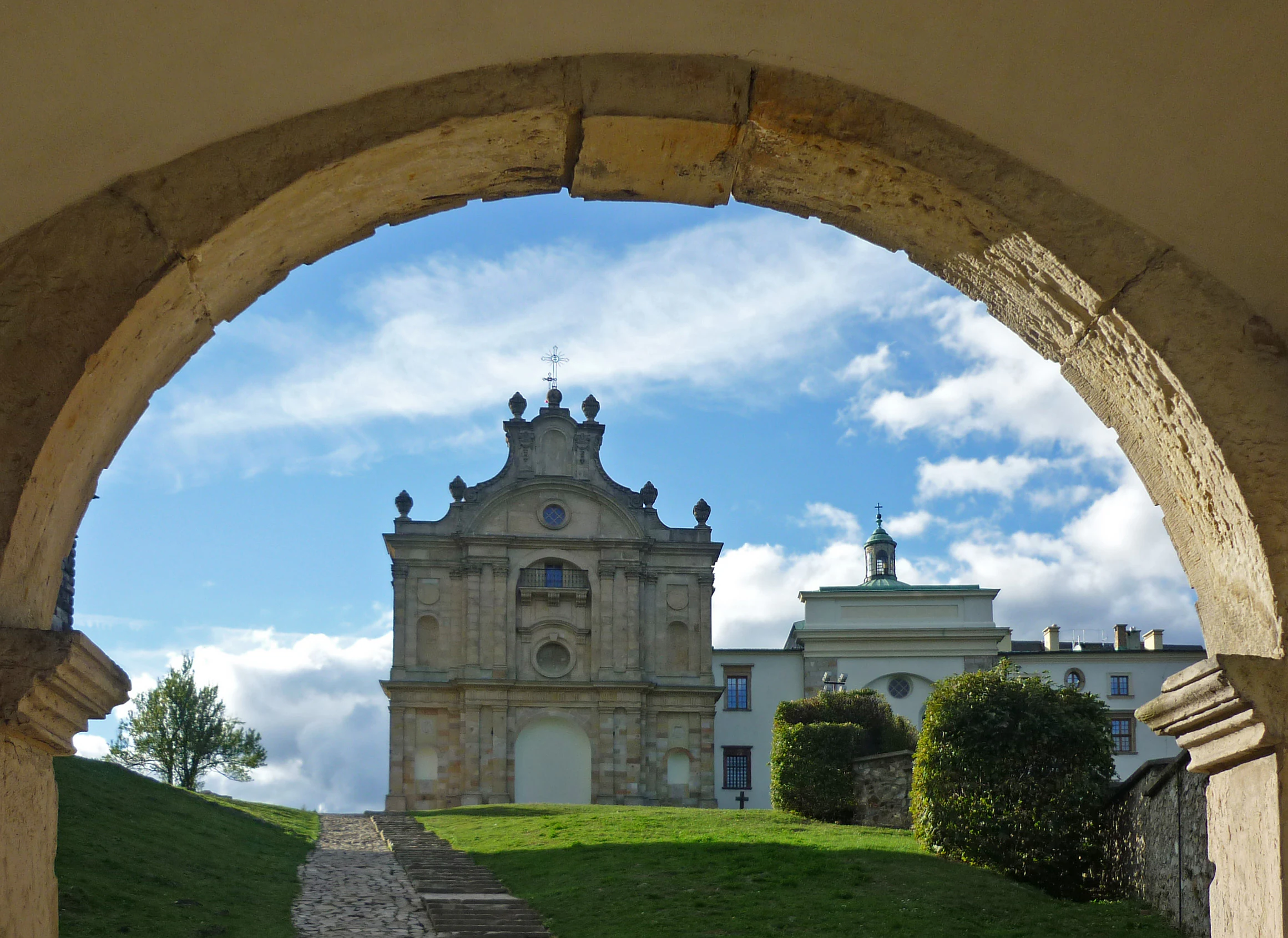 El Monasterio de la Santa Cruz en Polonia enmarcado a través de un gran arco de piedra con un sendero cubierto de hierba que conduce hacia el edificio.