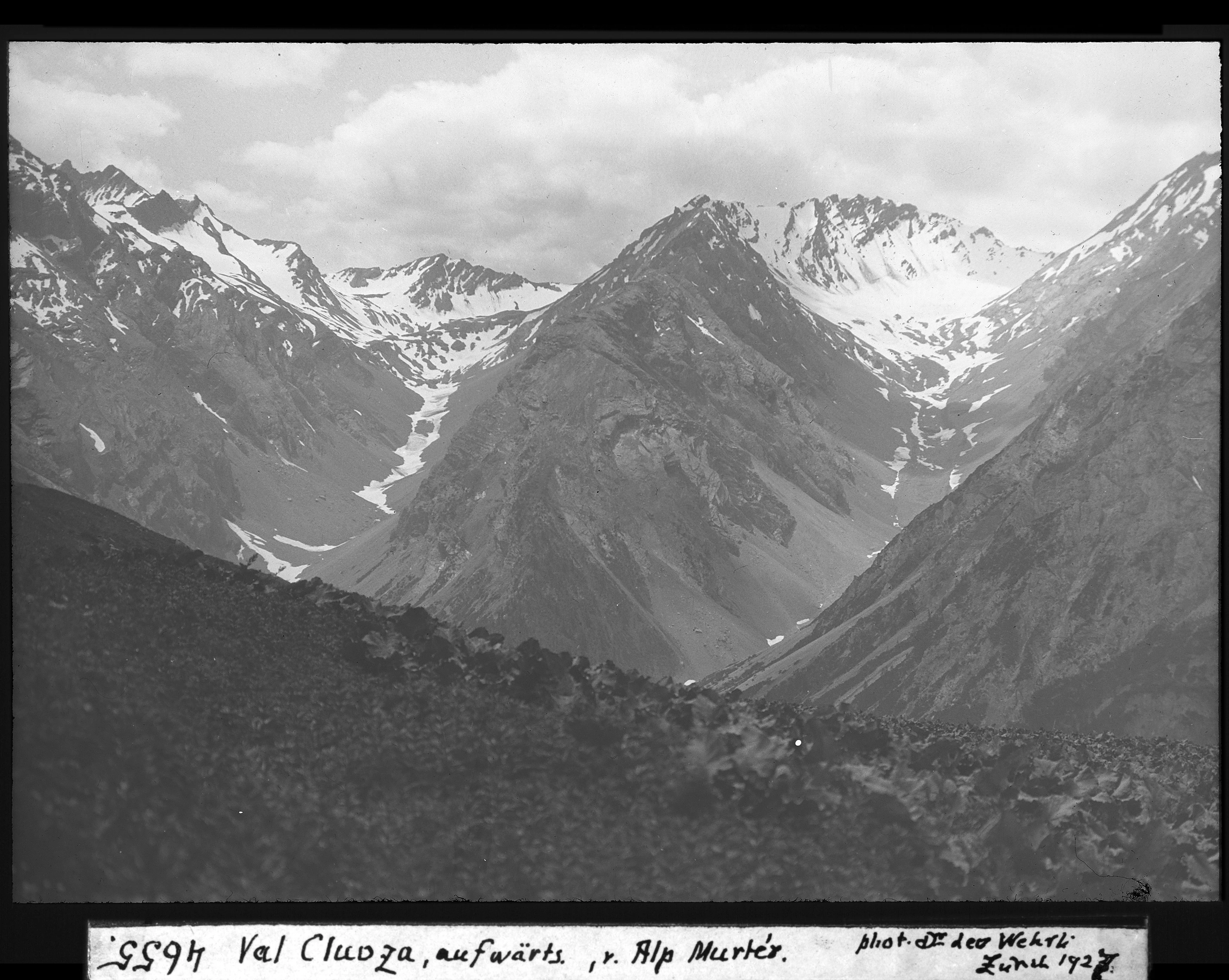 A black and white historical photograph of steep, snow-dusted alpine mountains rising over a deep valley, with a rocky, shadowed foreground.