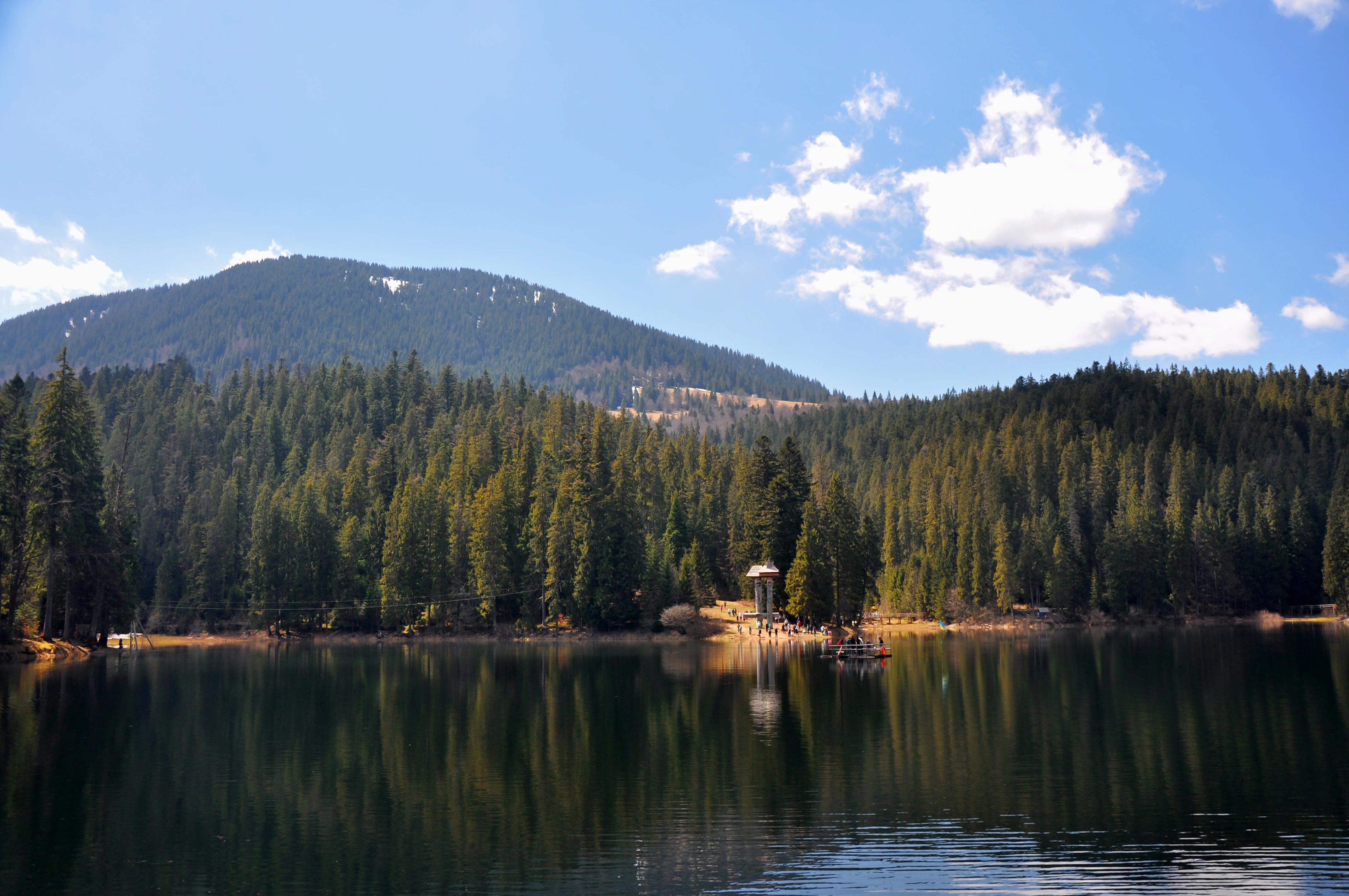 A calm lake reflecting dense evergreen forests and a mountain range under a partly cloudy sky