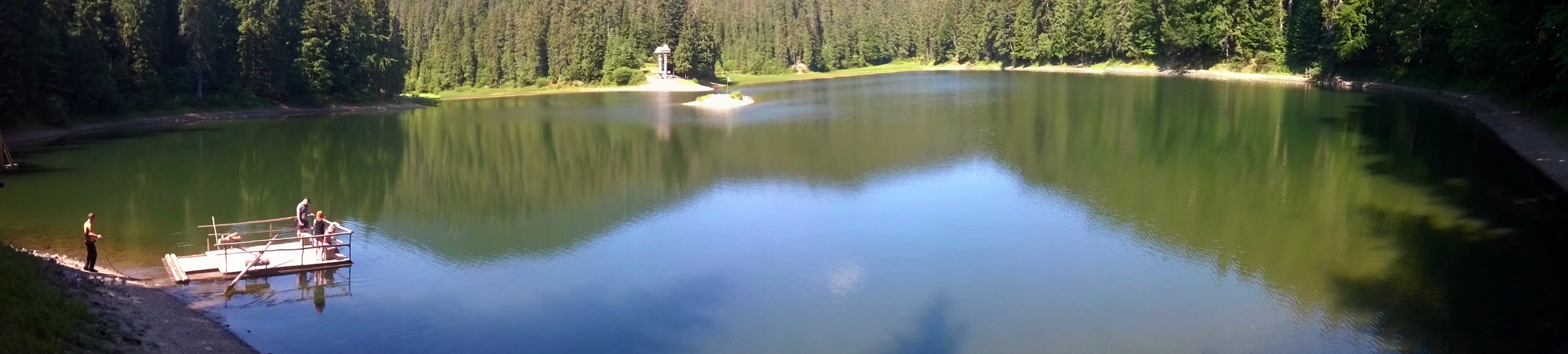 Panoramic view of Synevyr Lake with calm water reflecting surrounding dense forest, a small dock with two people on the left, and a small island in the distance