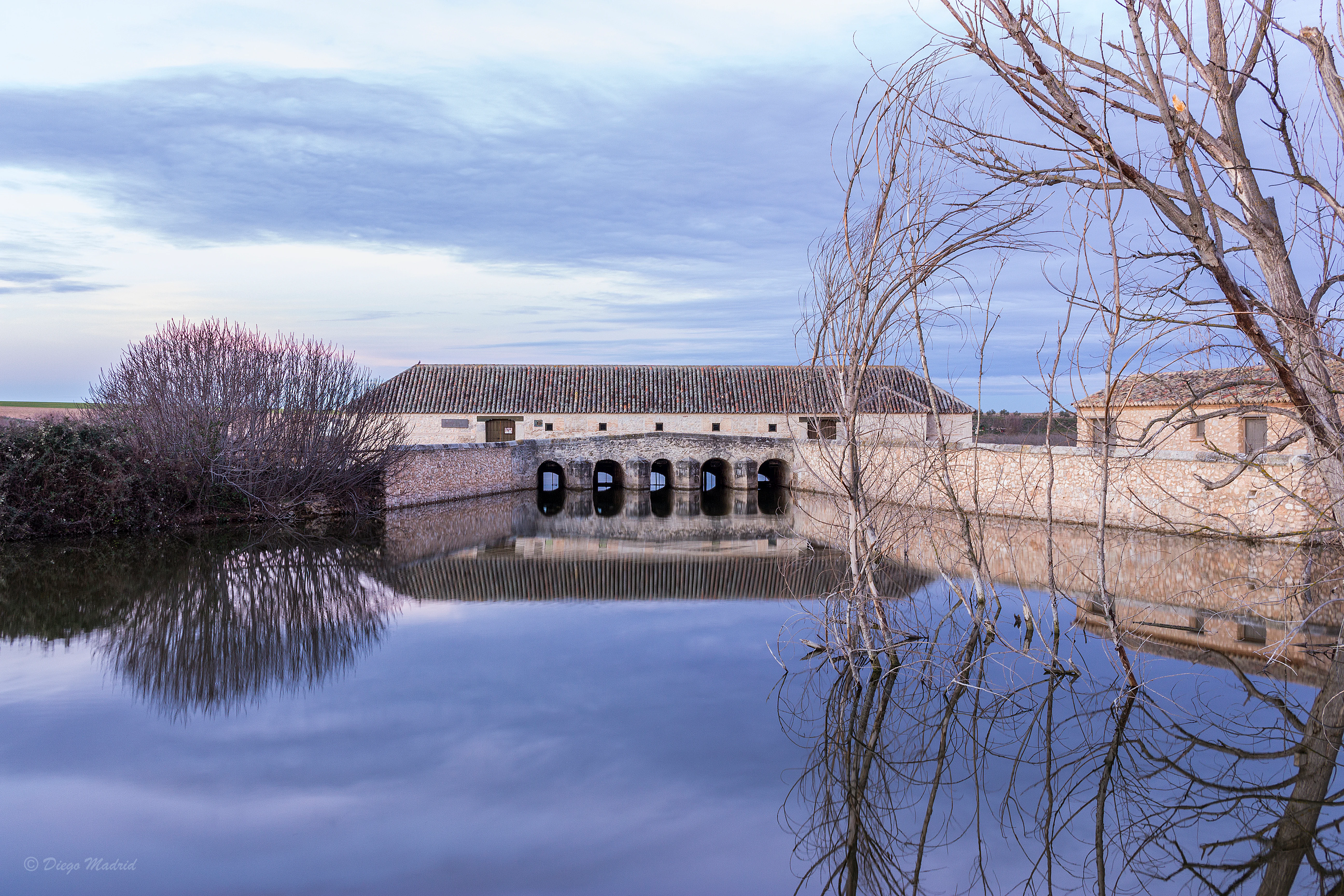Stone mill building with multiple arches reflected in still water, with bare trees and bushes nearby