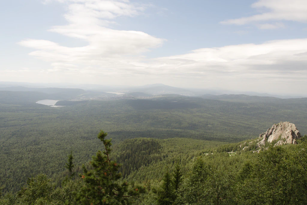 Panoramic landscape view of Taganay National Park showing forested mountains, a lake, and rocky outcrops under a partly cloudy sky