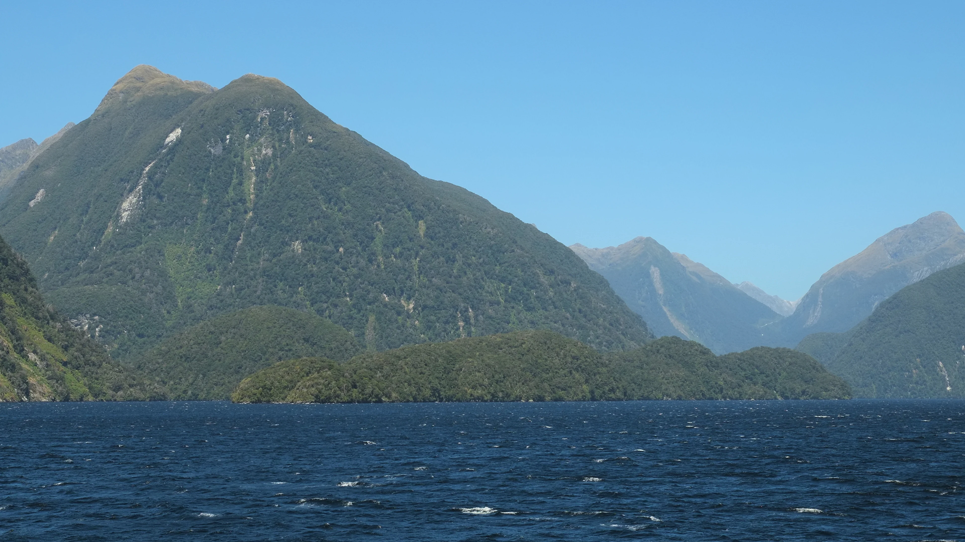 A wide view of a forest covered island surrounded by dark blue water, with steep mountains rising in the background under a clear sky.