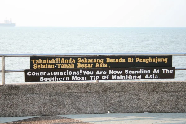 Un cartel con texto en malayo e inglés que indica que te encuentras en el extremo más meridional de la Asia continental, montado en una barandilla con vistas al mar.
