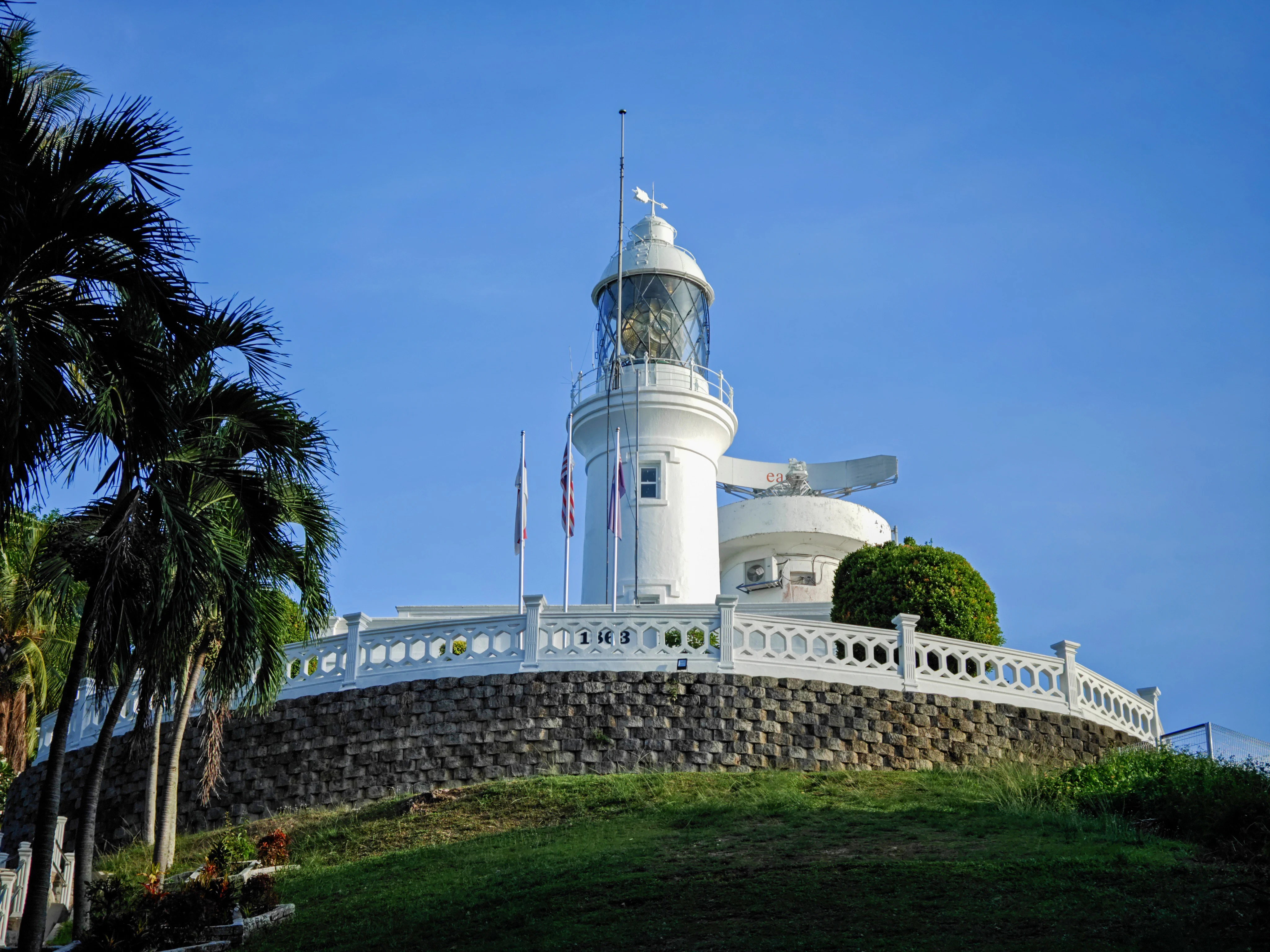 A white cylindrical lighthouse with a lantern room atop a stone base, surrounded by palm trees and a low white railing against a clear blue sky.