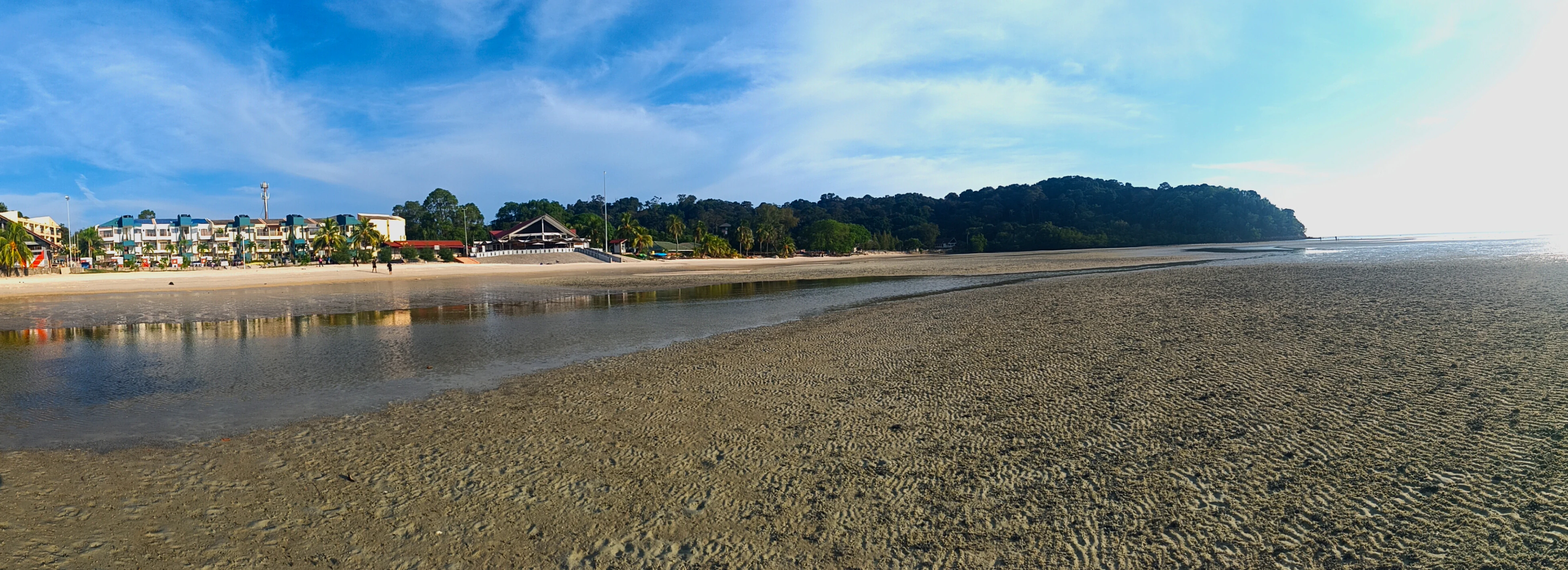 A panoramic photograph showing a wide, sandy beach at low tide with a reflective pool of water in the midground, backed by a treelined coast and buildings.