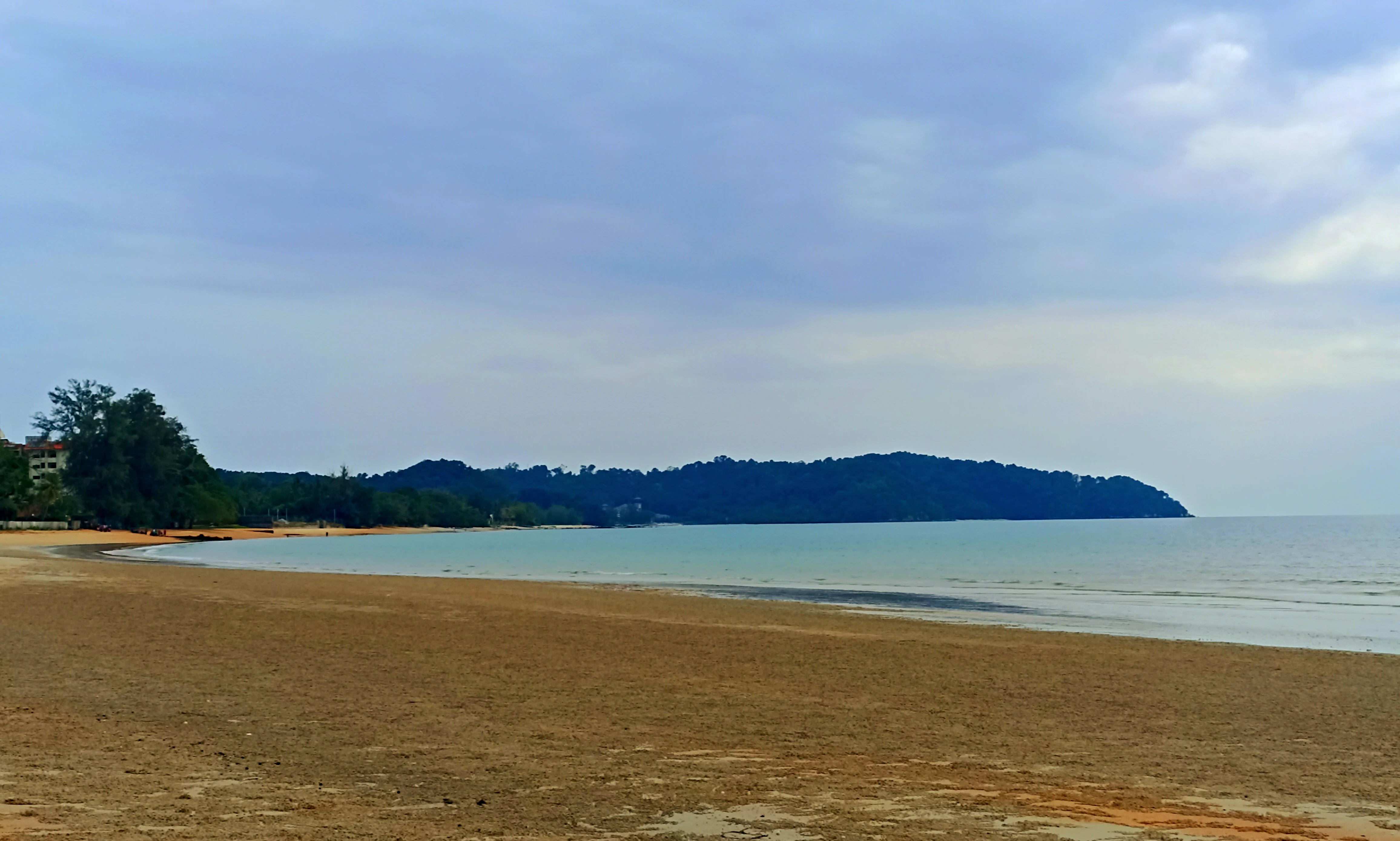 A wide view of a sandy beach in the foreground leading to calm water and a dark, forested cape in the distance under a cloudy sky.