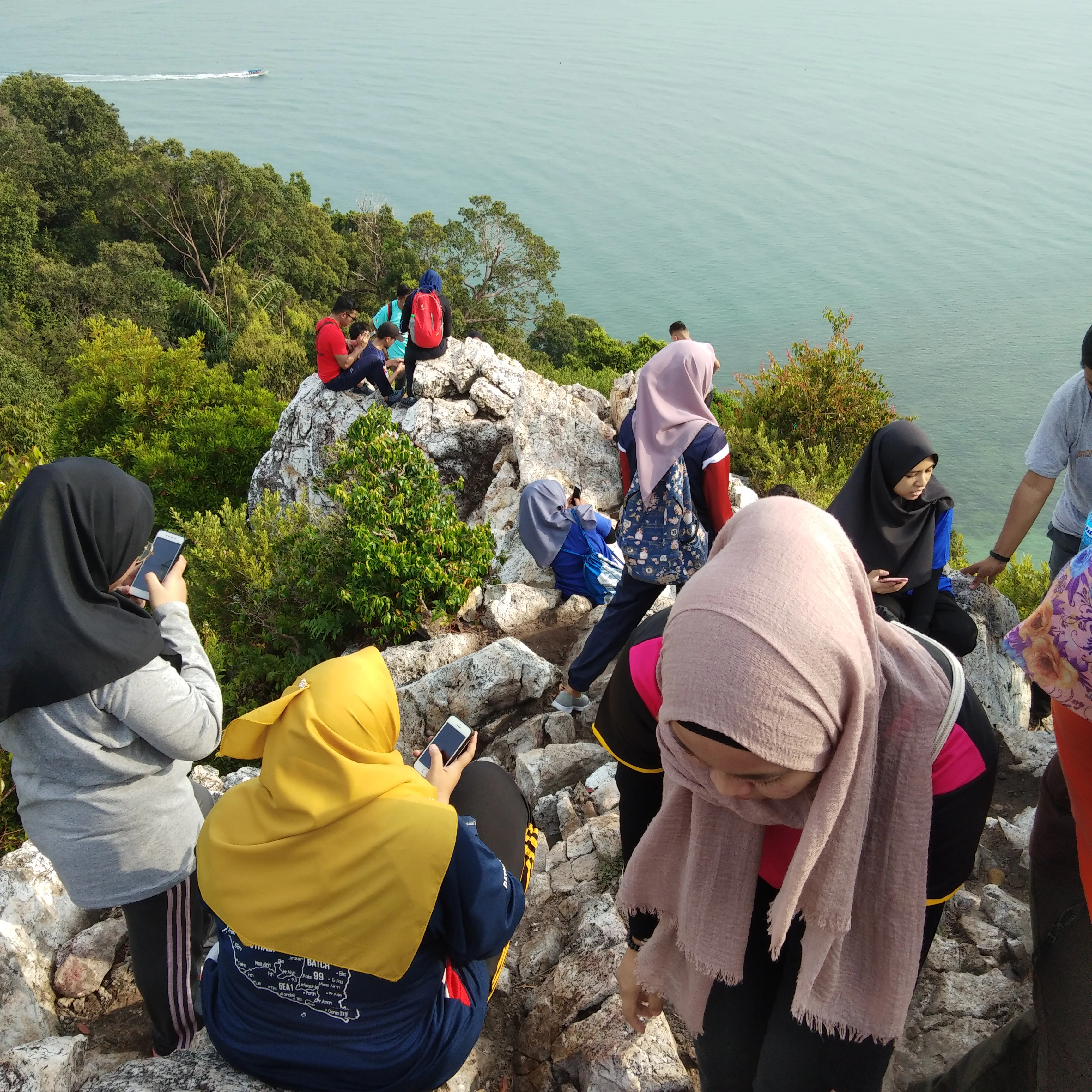 A group of people wearing hijabs and casual hiking clothes standing and sitting on a rocky, uneven summit overlooking a forested coastline and calm ocean.