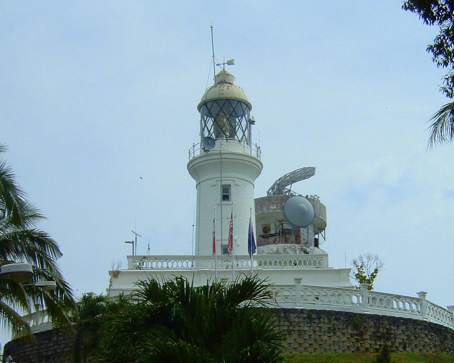 A white lighthouse with a lantern room and a nearby communications structure set atop a stone-walled elevation, framed by palm fronds under a clear sky.