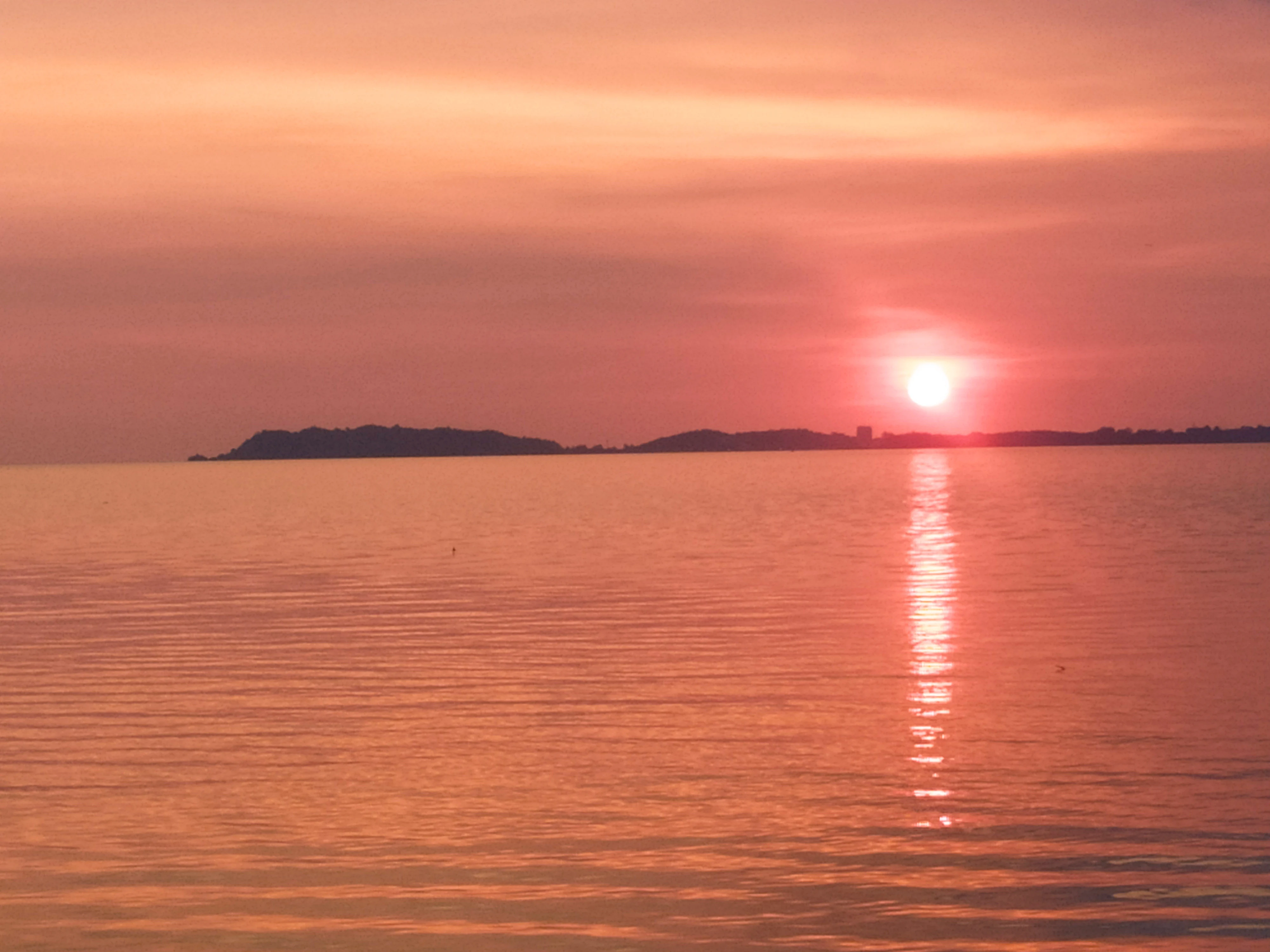 A vibrant sunset over a calm body of water with a dark silhouette of a coastal headland visible on the horizon.
