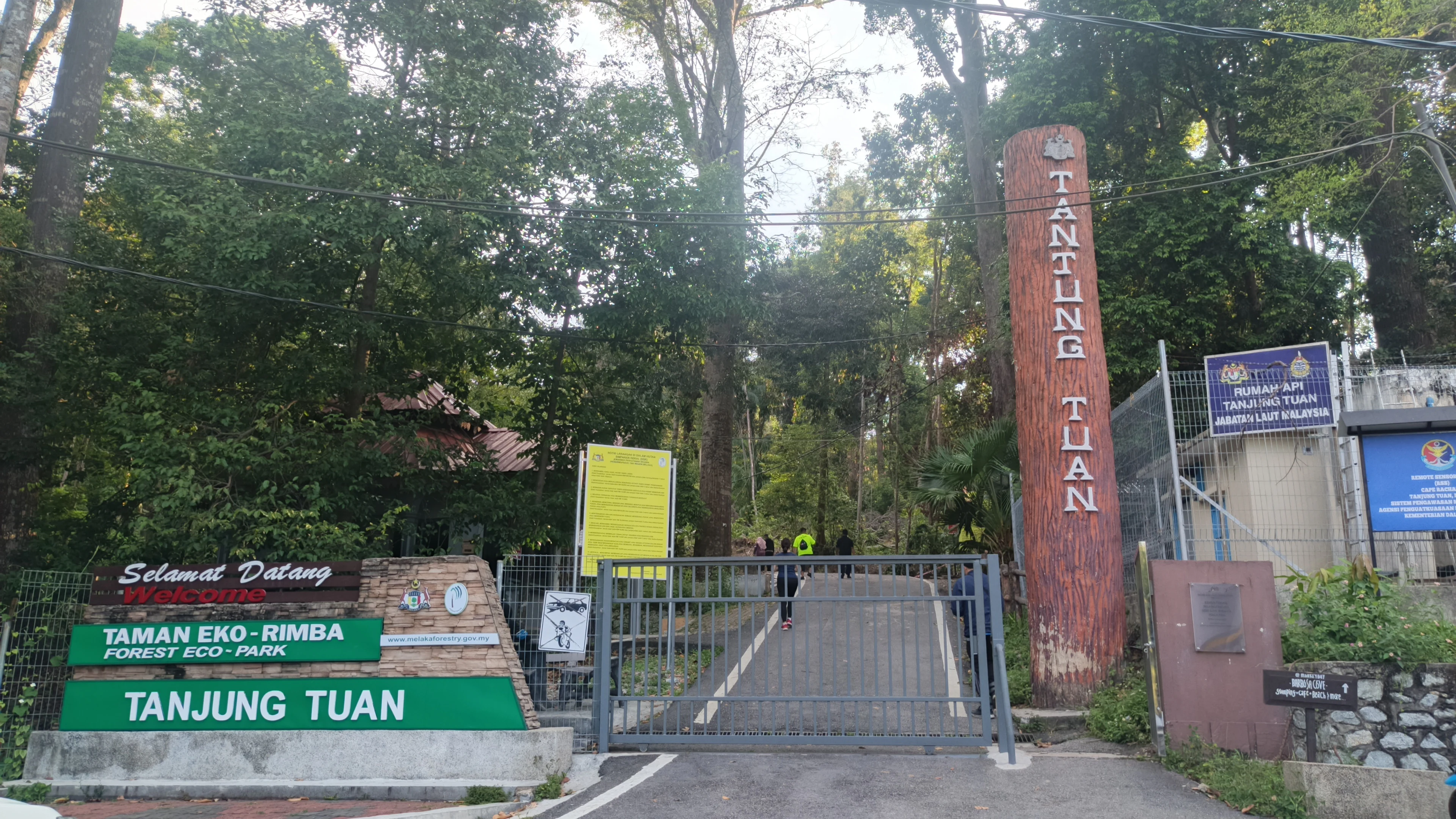 An entrance gate for Tanjung Tuan Forest Eco-Park, featuring green signs and a tall vertical log post with the name Tanjung Tuan engraved on it.