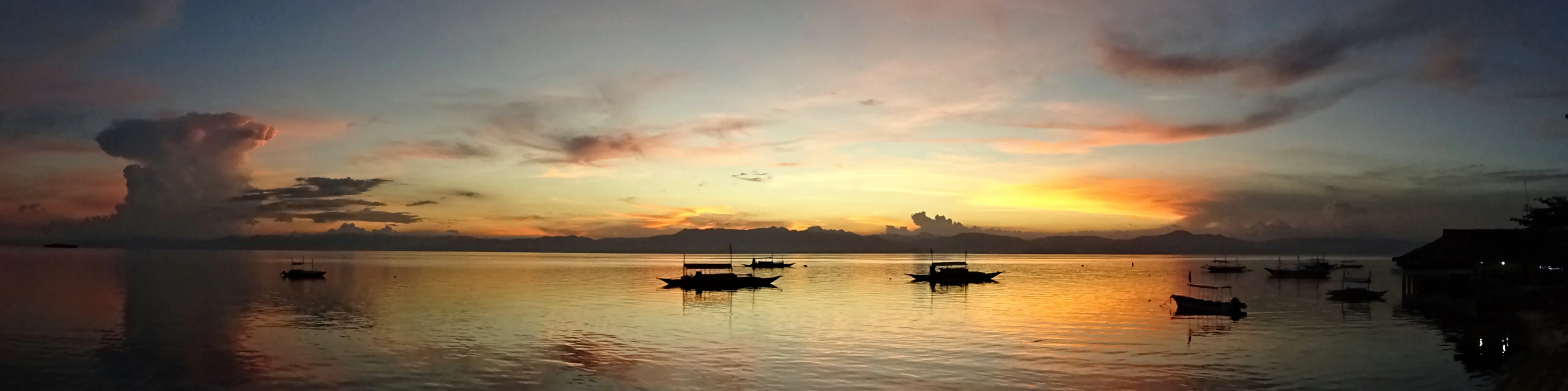 A wide panoramic view of a calm strait at sunset with small outrigger boats floating on the water, reflecting the orange and purple sky.