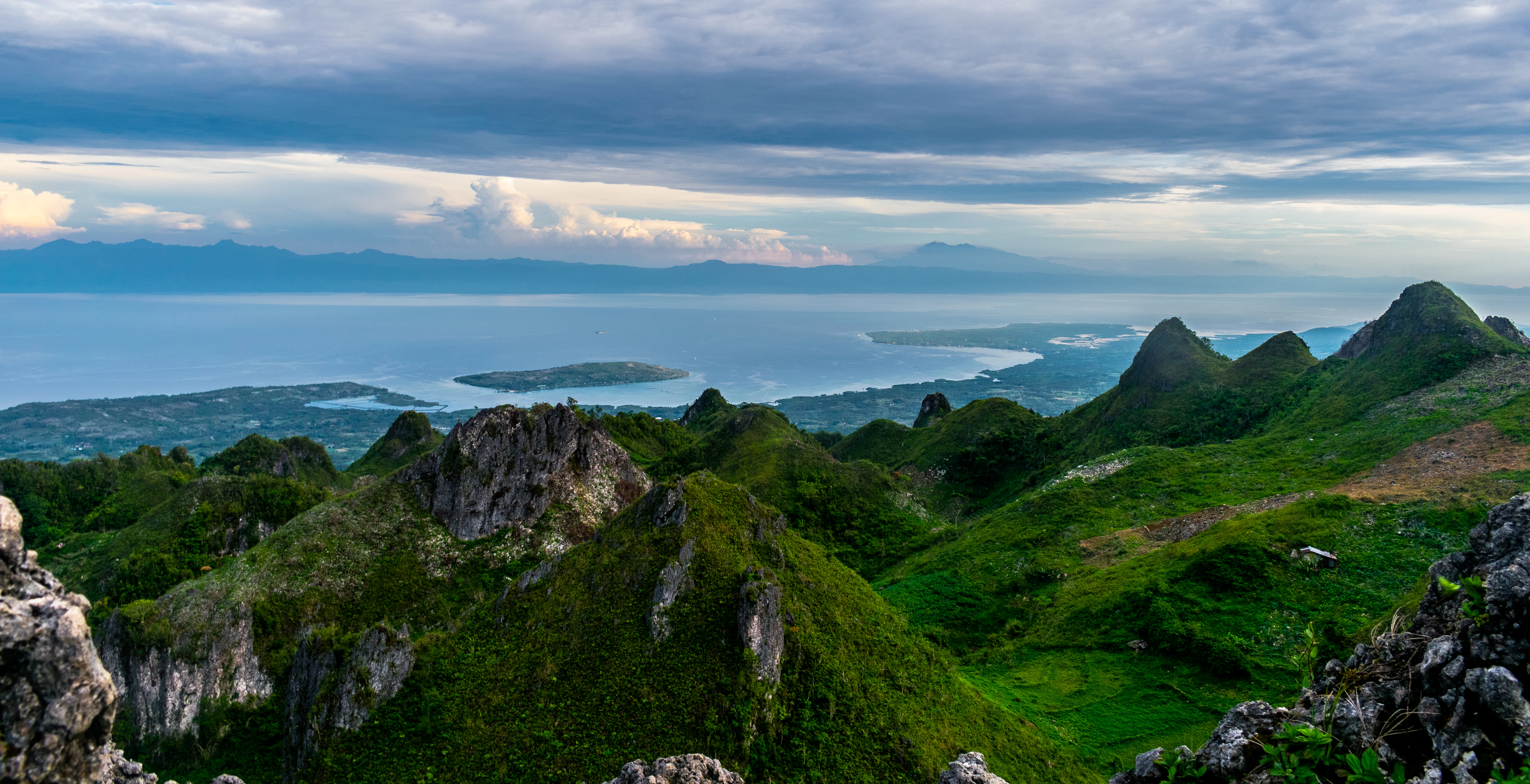 A high angle panoramic view of jagged, green karst hills in the foreground leading down to a calm, expansive strait of water with a distant island horizon under a cloudy sky.