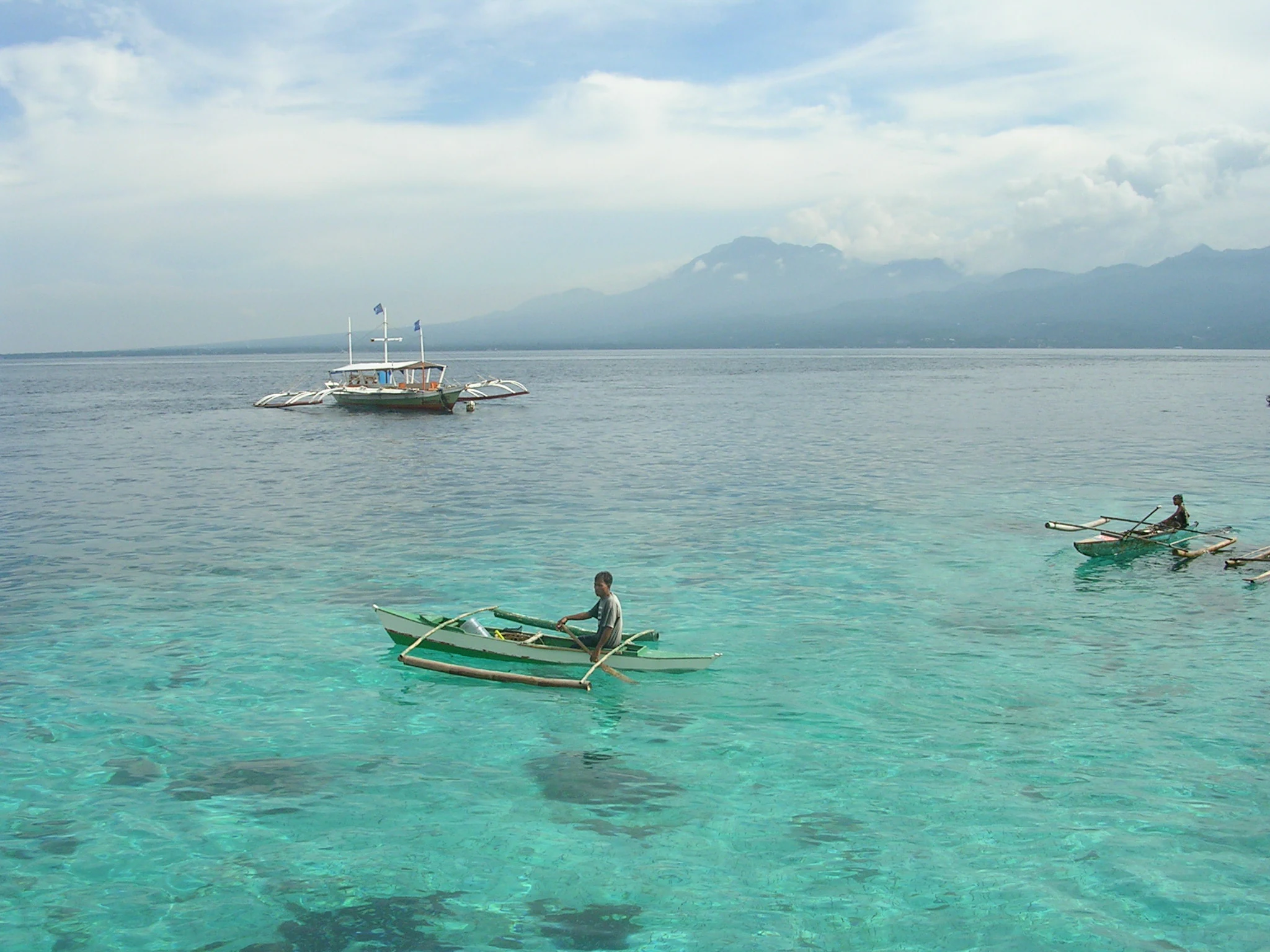 A person rowing a small wooden outrigger canoe in clear shallow turquoise water, with a larger boat and distant mountains visible across the strait.