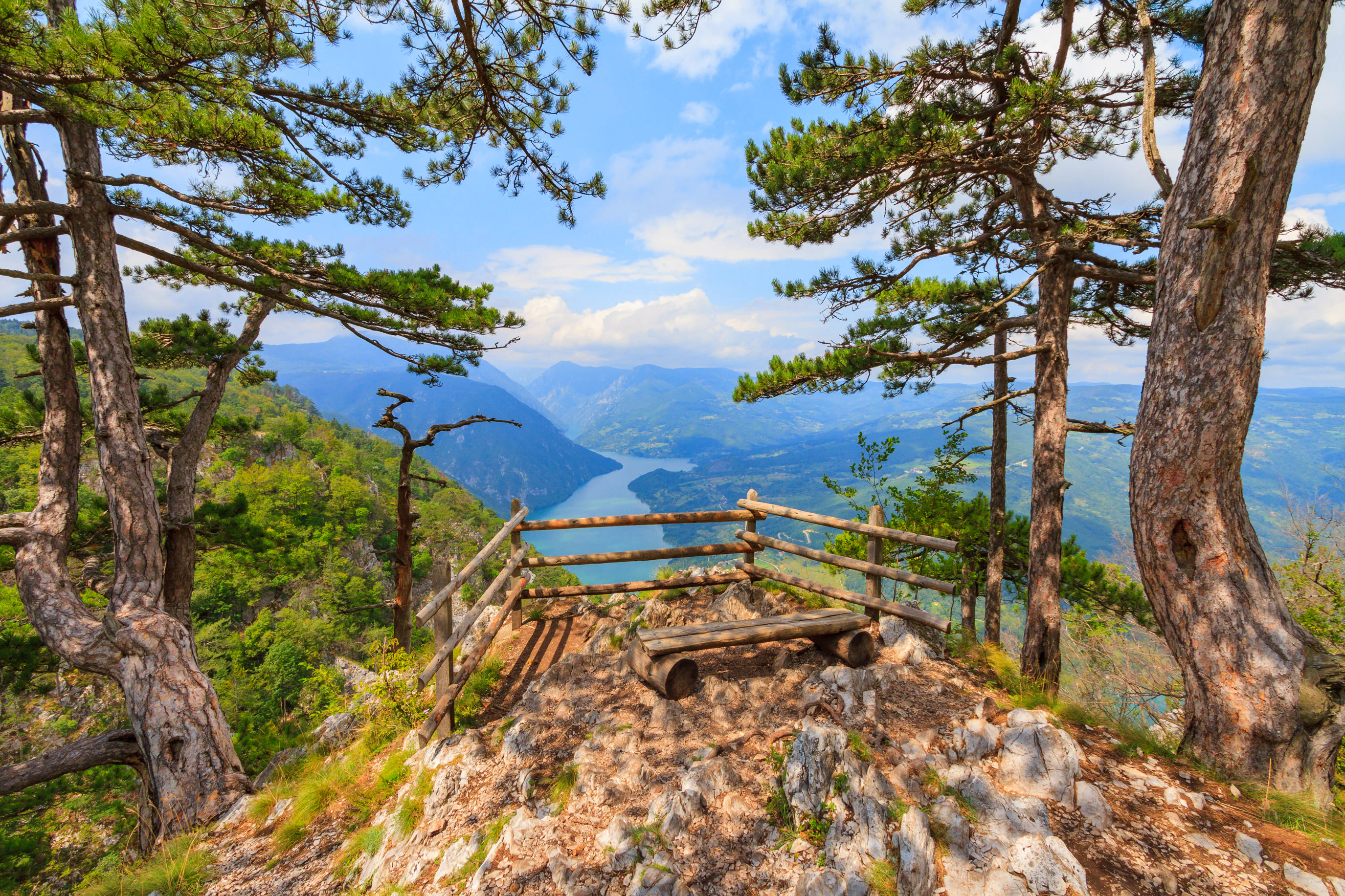 Wooden railing and bench at a mountain viewpoint overlooking a river valley with mountains and forest