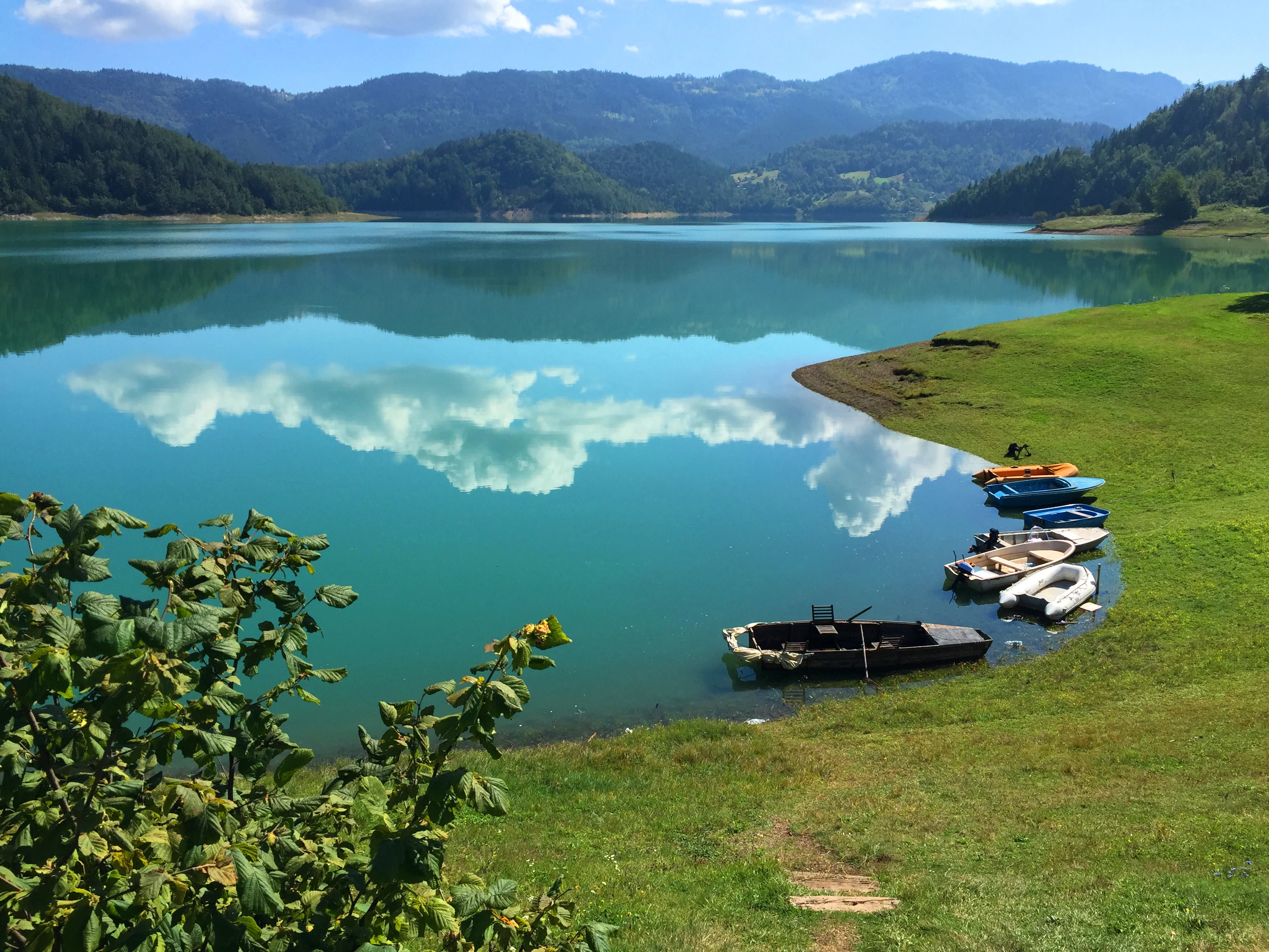 A scenic view of a turquoise lake reflecting clouds, with boats docked at a grassy shore and forested mountains in the background