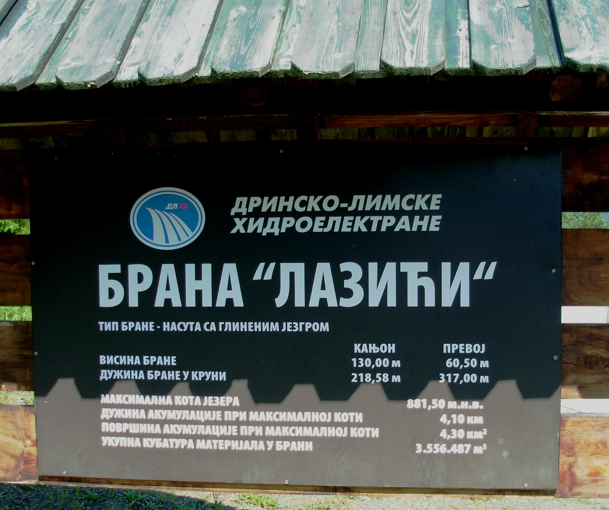 Black signboard with white text in Serbian Cyrillic script mounted on wooden structure, displaying technical specifications of Lazići Dam including height and volume measurements