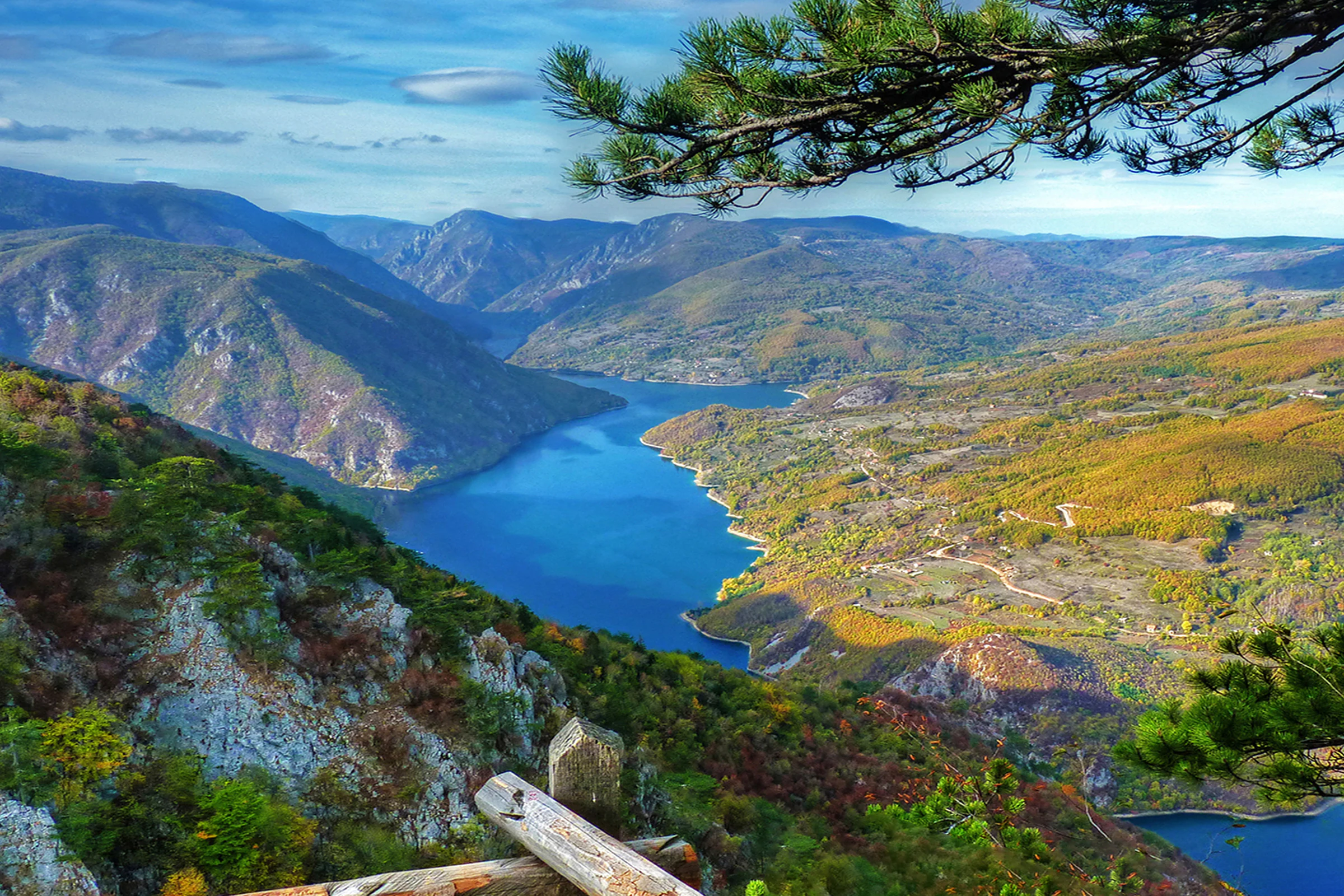Panoramic view of a river valley with mountains, forests, and a wooden railing in the foreground
