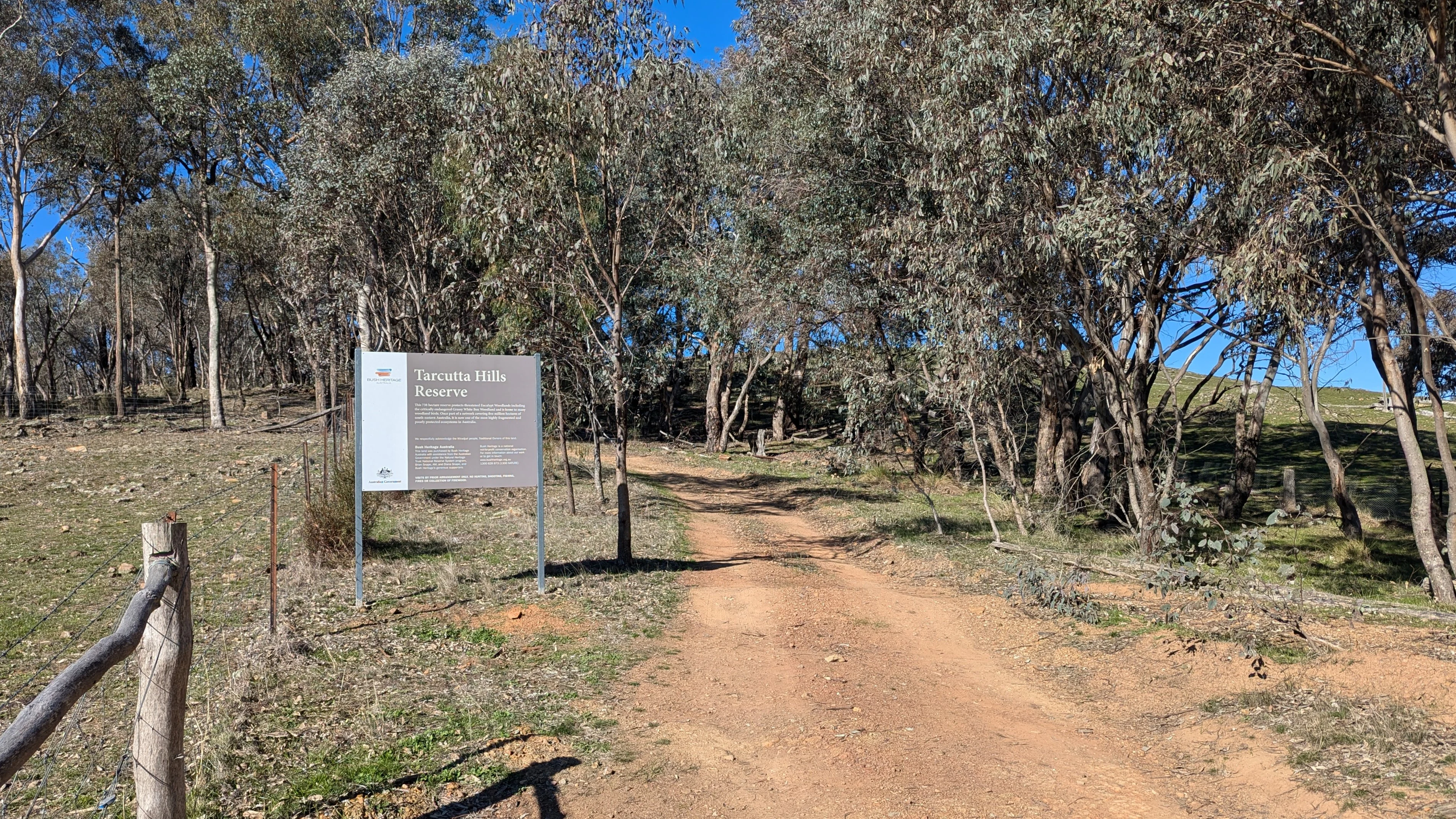 A dirt access road leads into a woodland area at Tarcutta Hills Reserve, with an information sign on the left side of the frame.