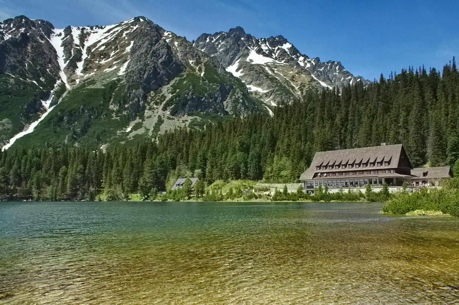 Lago claro rodeado de bosque denso con montañas nevadas y un edificio de albergue