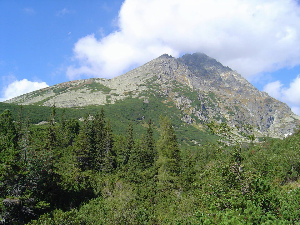 Pico montañoso con laderas rocosas y vegetación verde, rodeado de zonas boscosas bajo un cielo parcialmente nublado