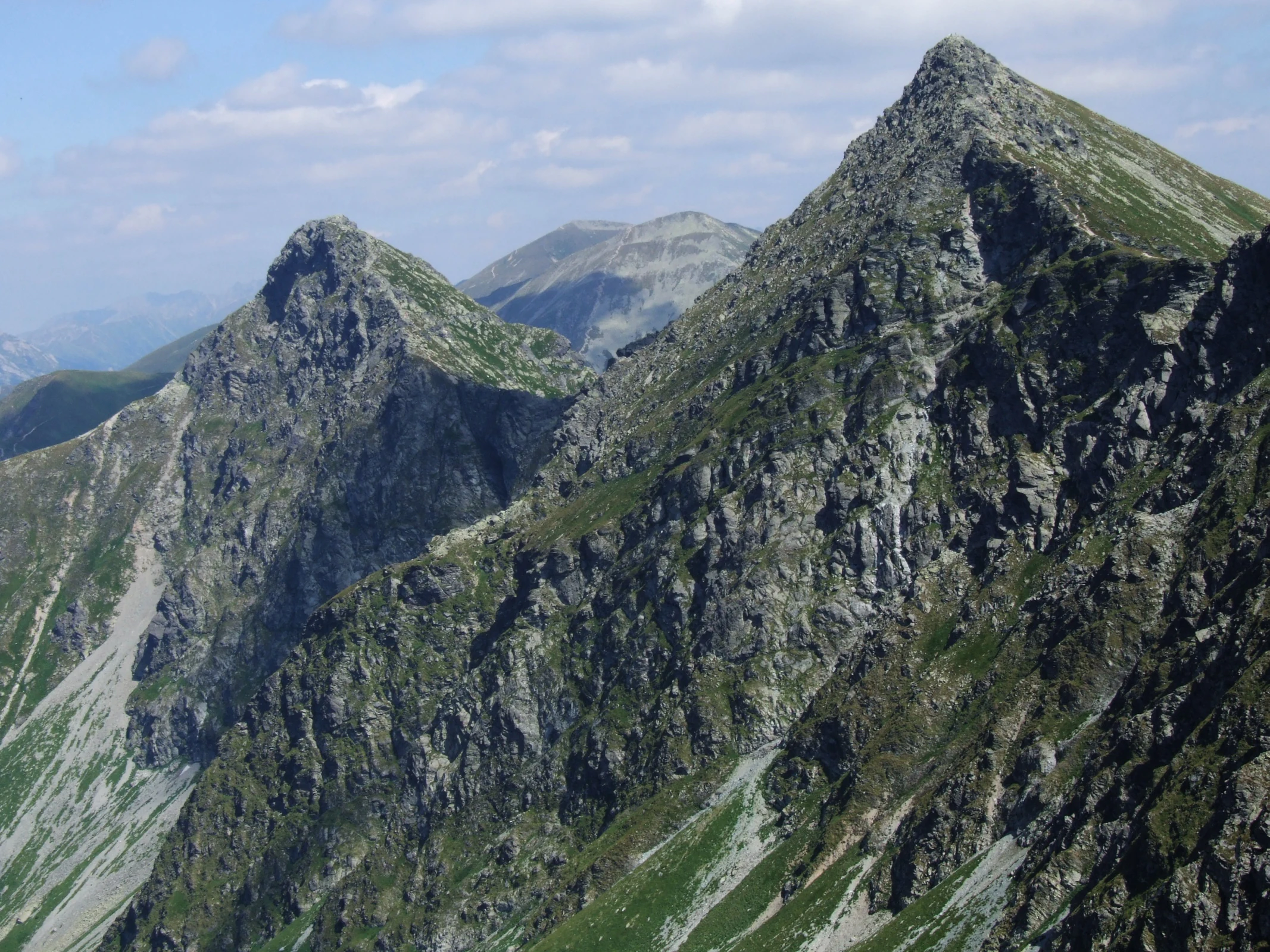 Picos montañosos escarpados con caras rocosas y parches de vegetación verde bajo un cielo parcialmente nublado