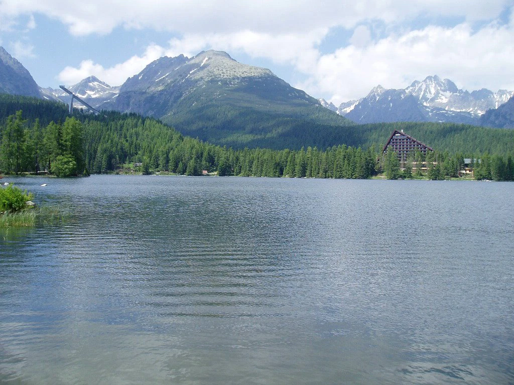 Aguas tranquilas de lago reflejando montañas distantes con cumbres nevadas bajo un cielo parcialmente nublado, rodeado por un denso bosque de coníferas con una gran cabaña de madera visible en la orilla derecha