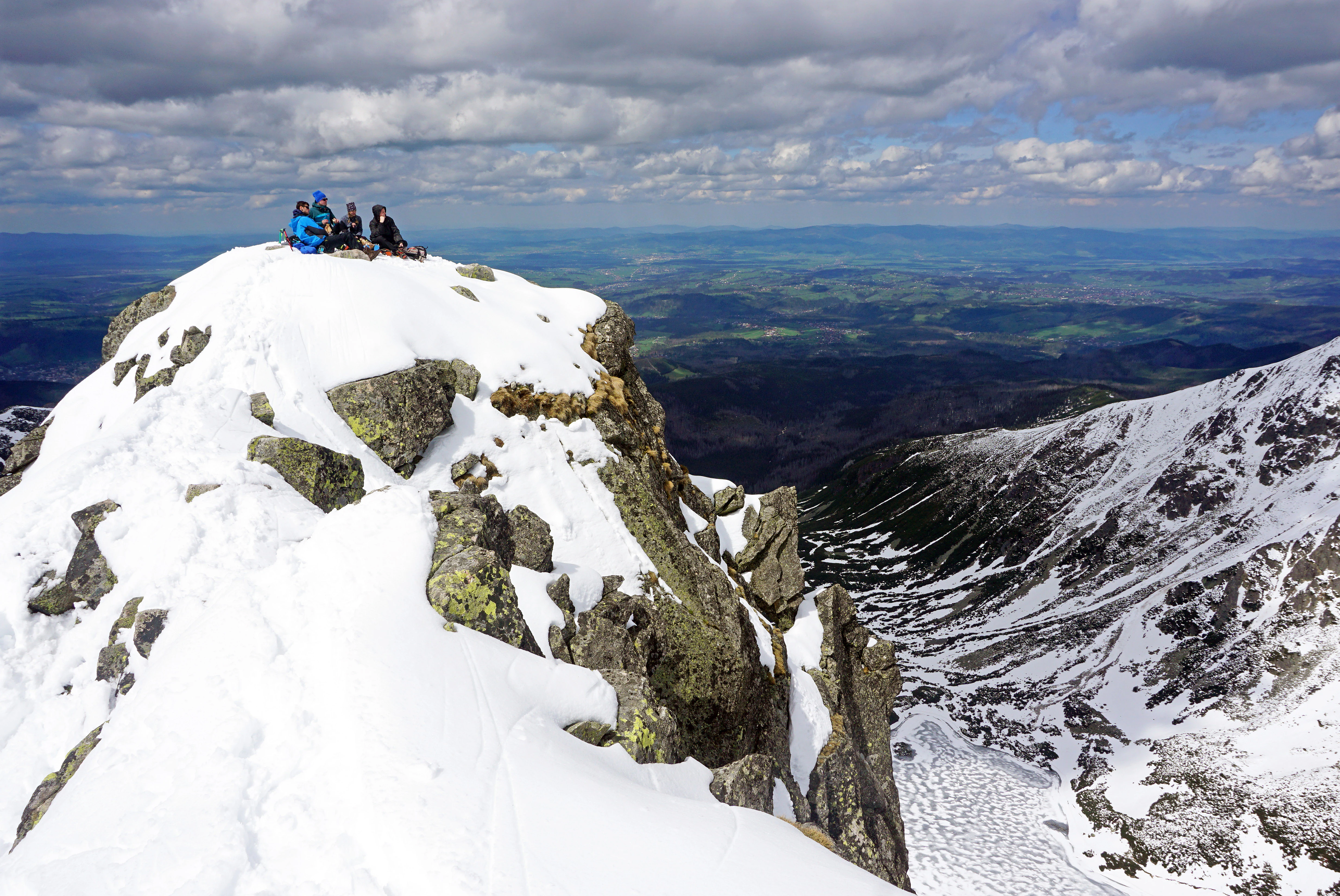 Four hikers sitting on a snow-covered mountain peak with rocky outcrops and a valley view in the background