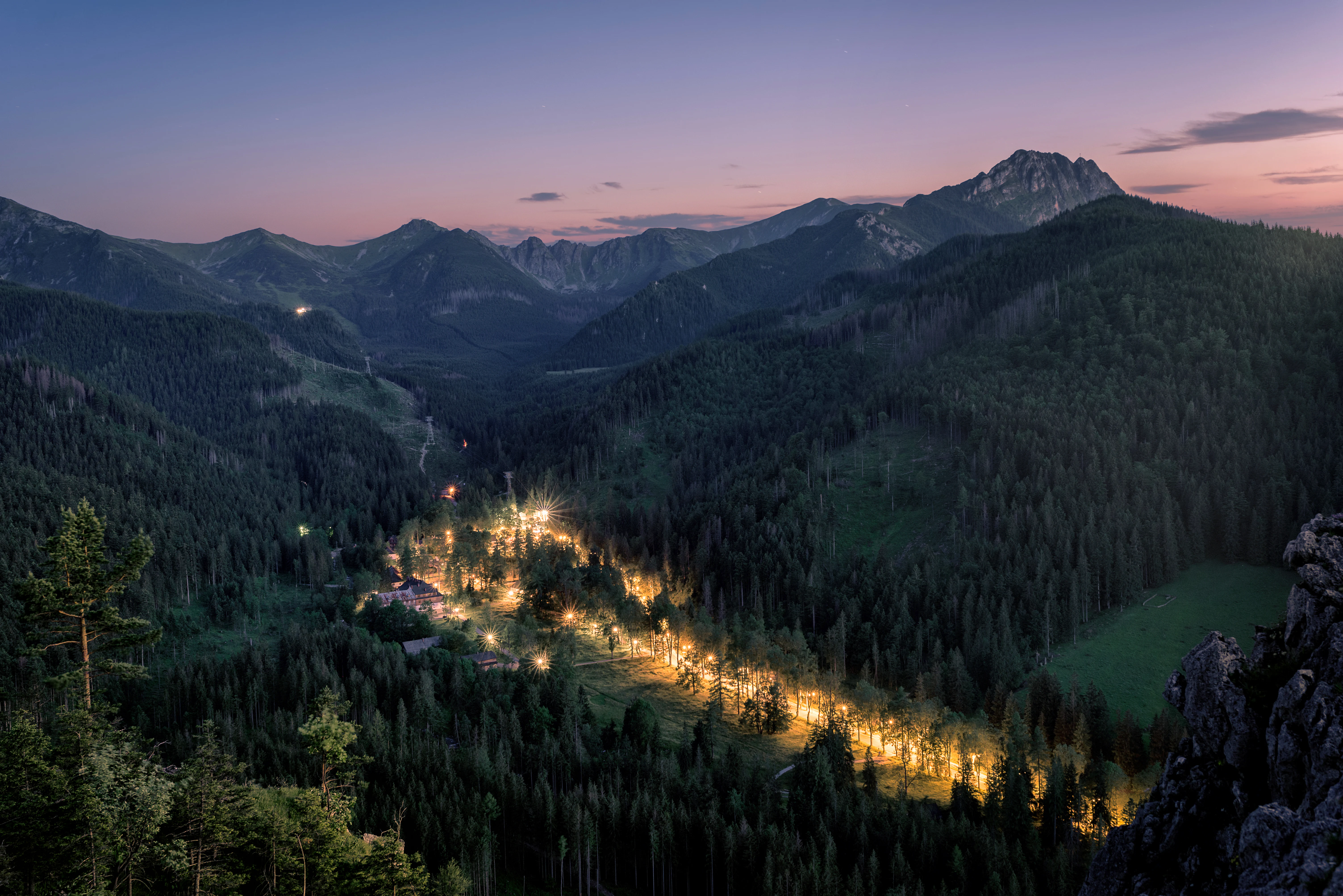 Mountain landscape at dusk with forested hills, illuminated valley path, and mountain peaks