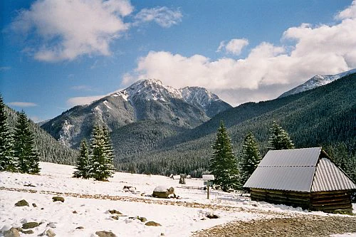 Snow-covered valley with a wooden cabin, pine trees, and snow-capped mountains under a partly cloudy sky