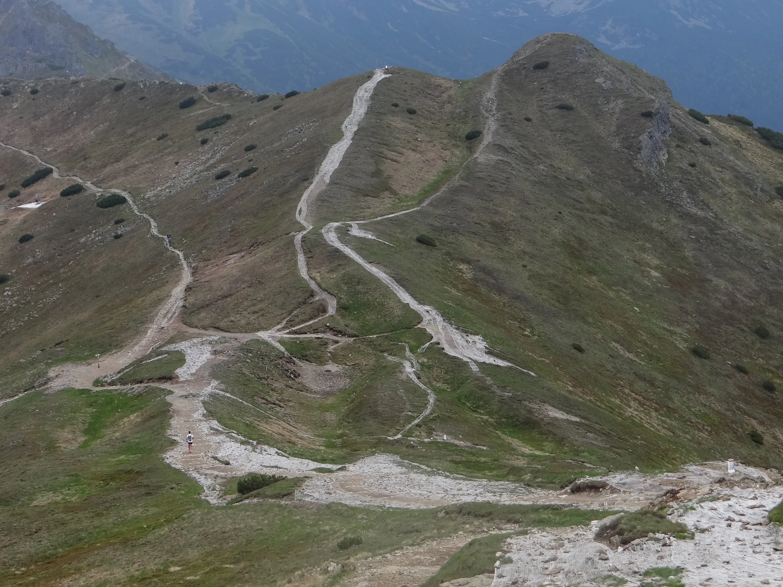 Una vista elevada en picado hacia un paso de montaña en los montes Tatra, con múltiples senderos de grava desgastados que atraviesan laderas alpinas verdes y marrones.