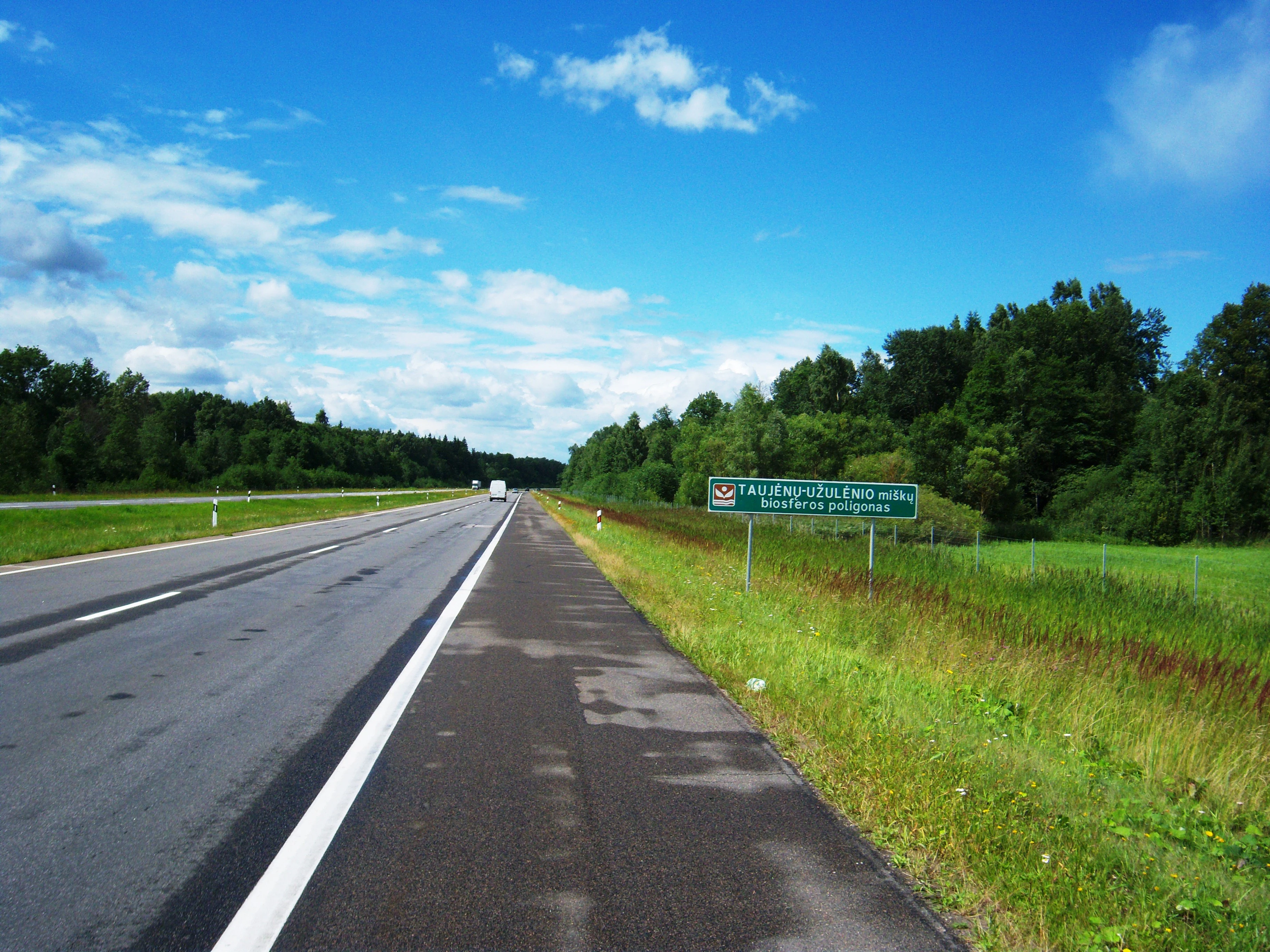 A view of a two-lane asphalt road stretching into the distance with a green park entrance sign on the right shoulder, surrounded by a grassy verge and dense forest.