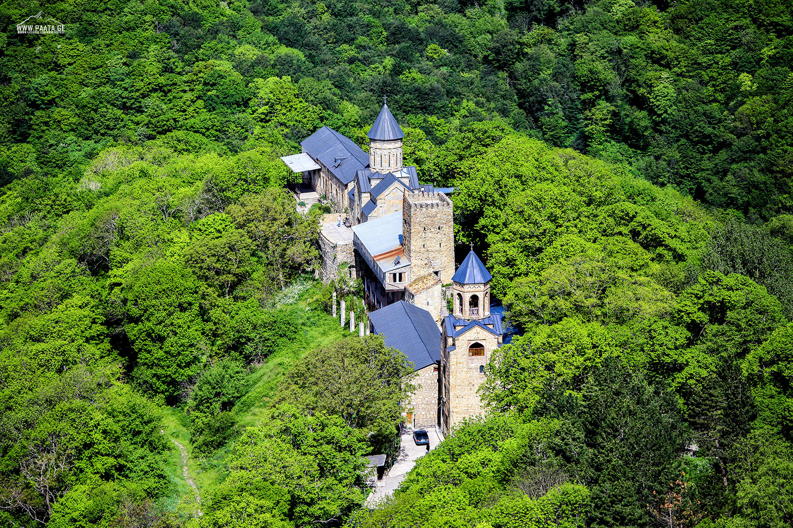 Aerial view of stone building complex with blue roofs surrounded by dense green forest on a hillside