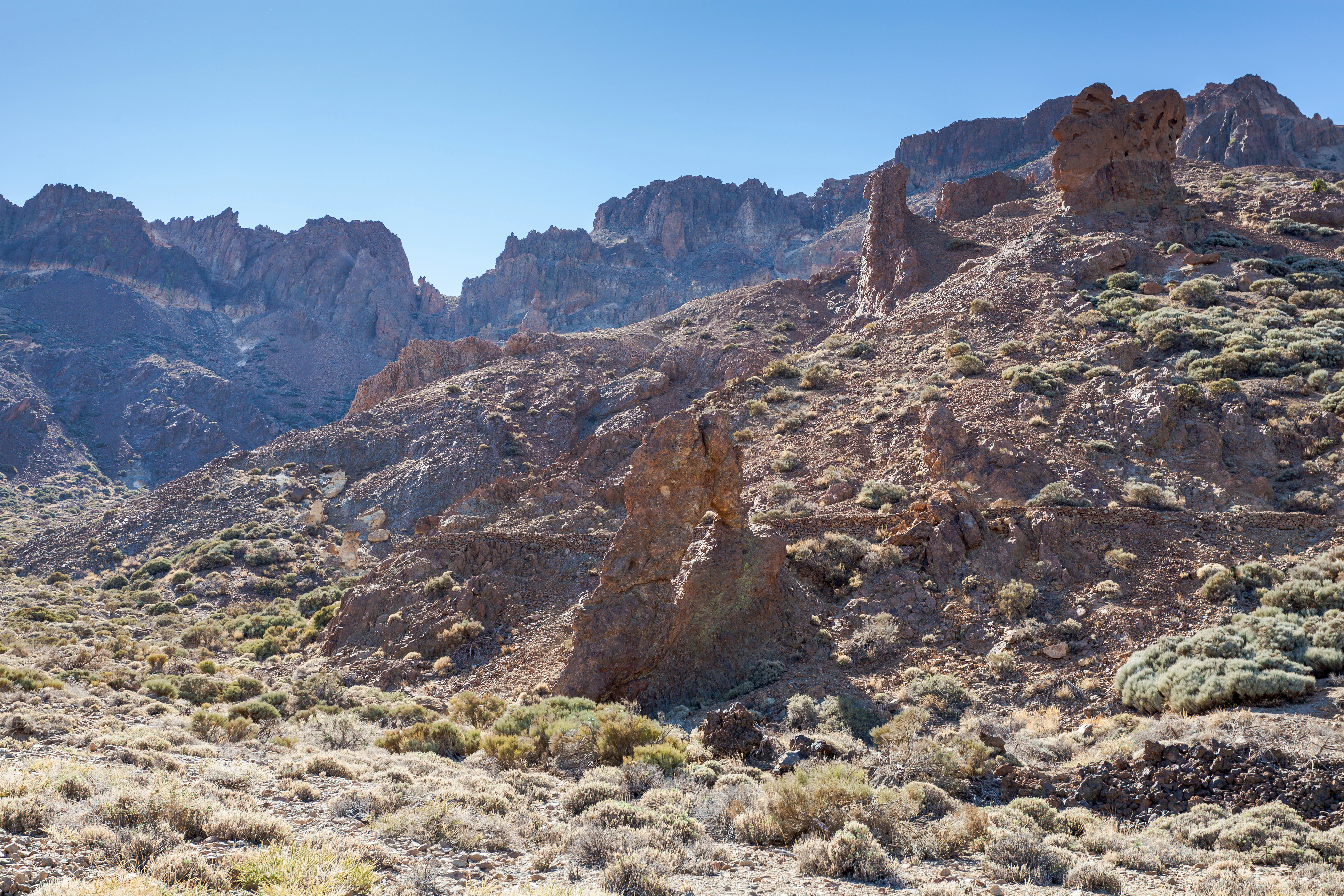 Volcanic landscape with rocky formations, sparse vegetation, and mountainous terrain under clear blue sky