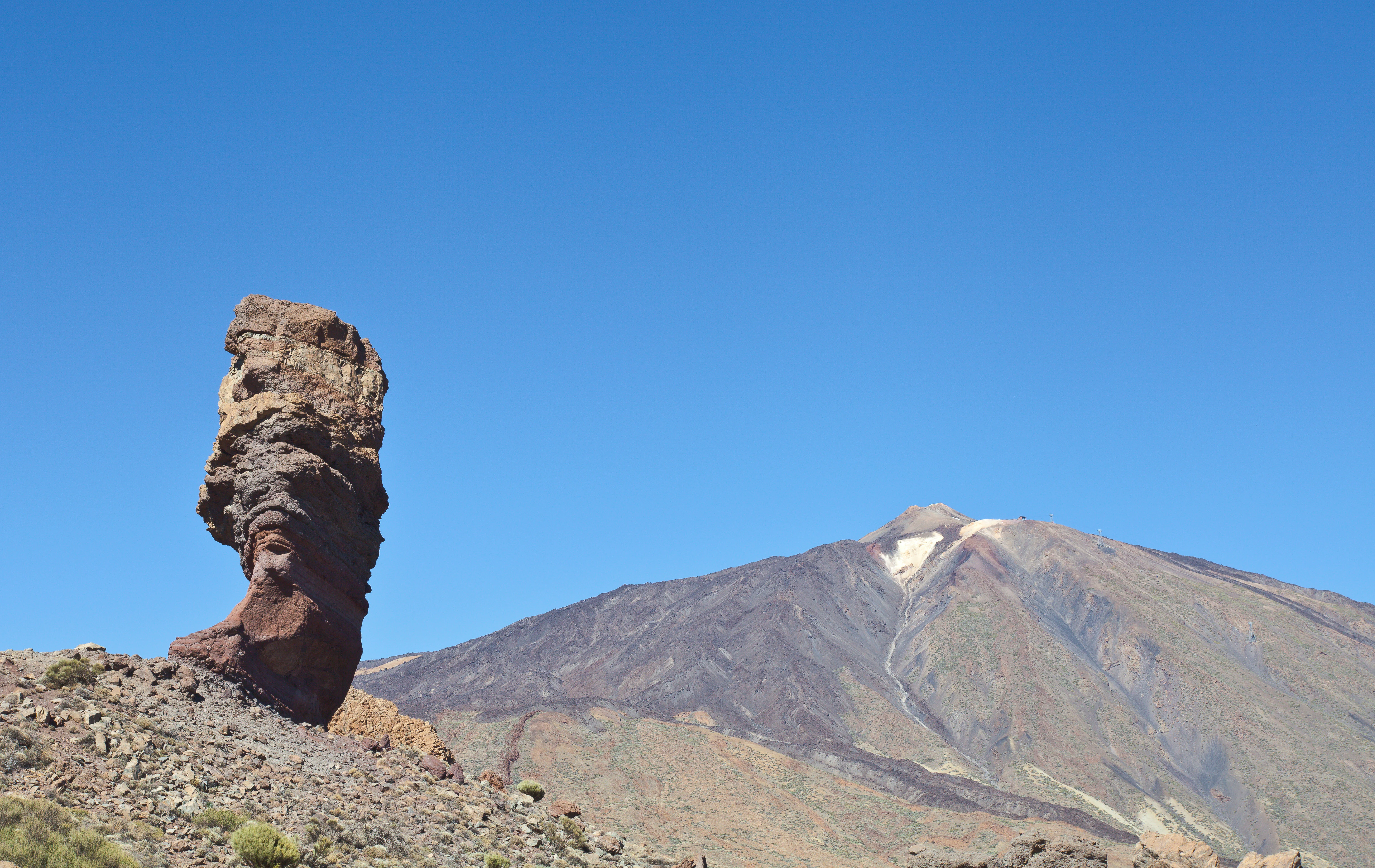 Tall weathered rock formation (Roque Cinchado) on rocky terrain with Mount Teide mountain visible under clear blue sky