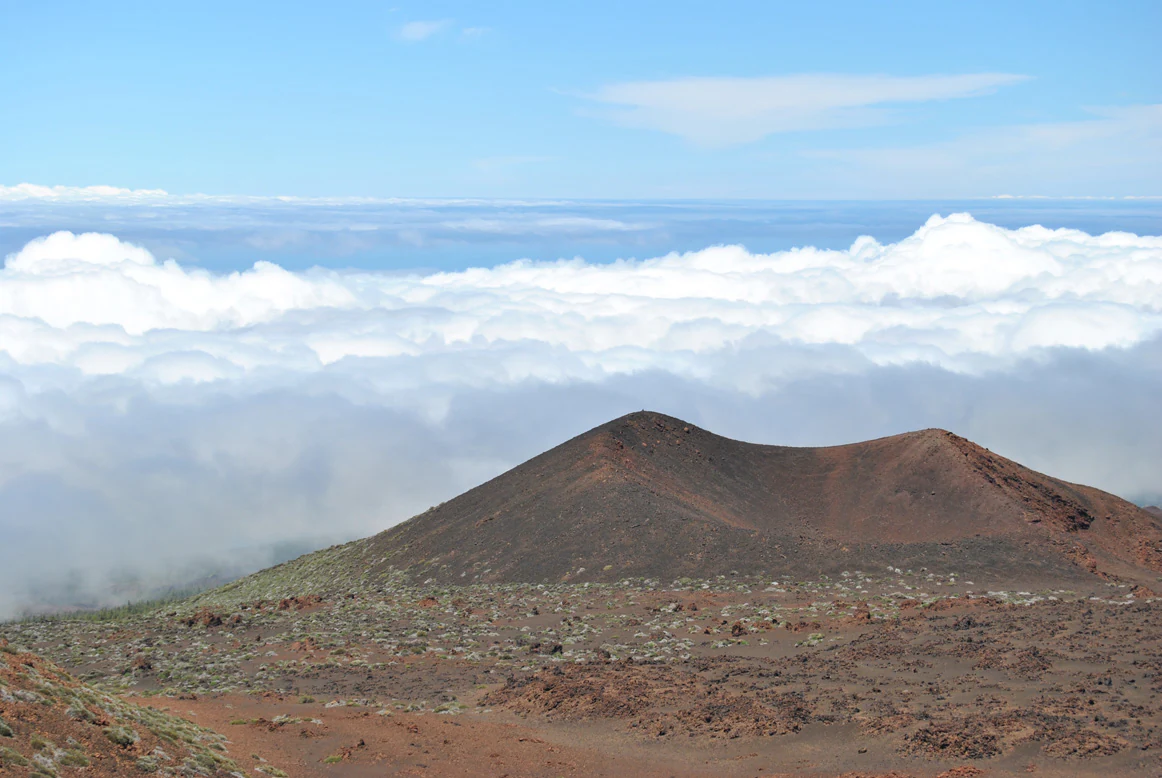 A volcanic mountain peak rises above a layer of clouds with clear blue sky above, showing the distinctive reddish-brown terrain of Teide National Park