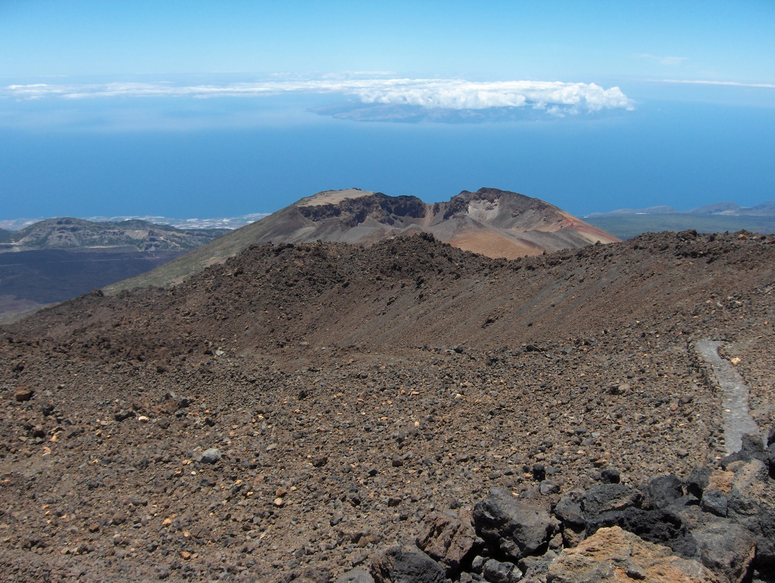 Volcanic crater landscape with rocky terrain, a trail on the right, and distant ocean with La Gomera island visible