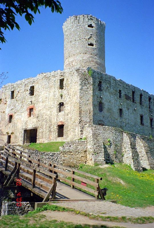 A low-angle view of the weathered stone ruins of Lipowiec Castle, featuring a prominent circular tower and an adjacent wooden footbridge leading to the castle entrance.
