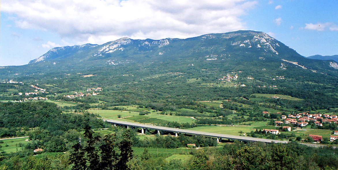 A wide mountain landscape showing a large, forested mountain ridge in the background, with a valley containing a highway viaduct and small villages in the foreground.