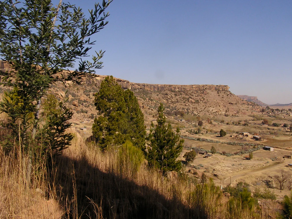 A wide view of a dry, arid landscape showing the prominent Thaba Bosiu sandstone plateau rising above a valley with scattered vegetation and small structures.