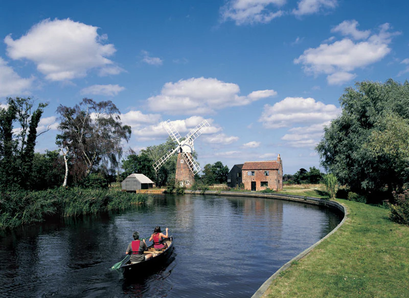 A river with a boat carrying two people, a watermill and buildings in the background, surrounded by trees under a blue sky with clouds
