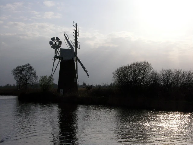 Windmill silhouette on a river with trees and a partly cloudy sky